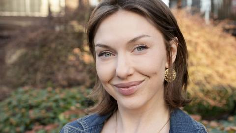 Woman with short auburn hair and gold earrings with fall vegetation in background.