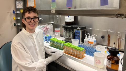 Person with short curly brown hair and glasses sits in a lab near a counter full of test tubes and other equipment.