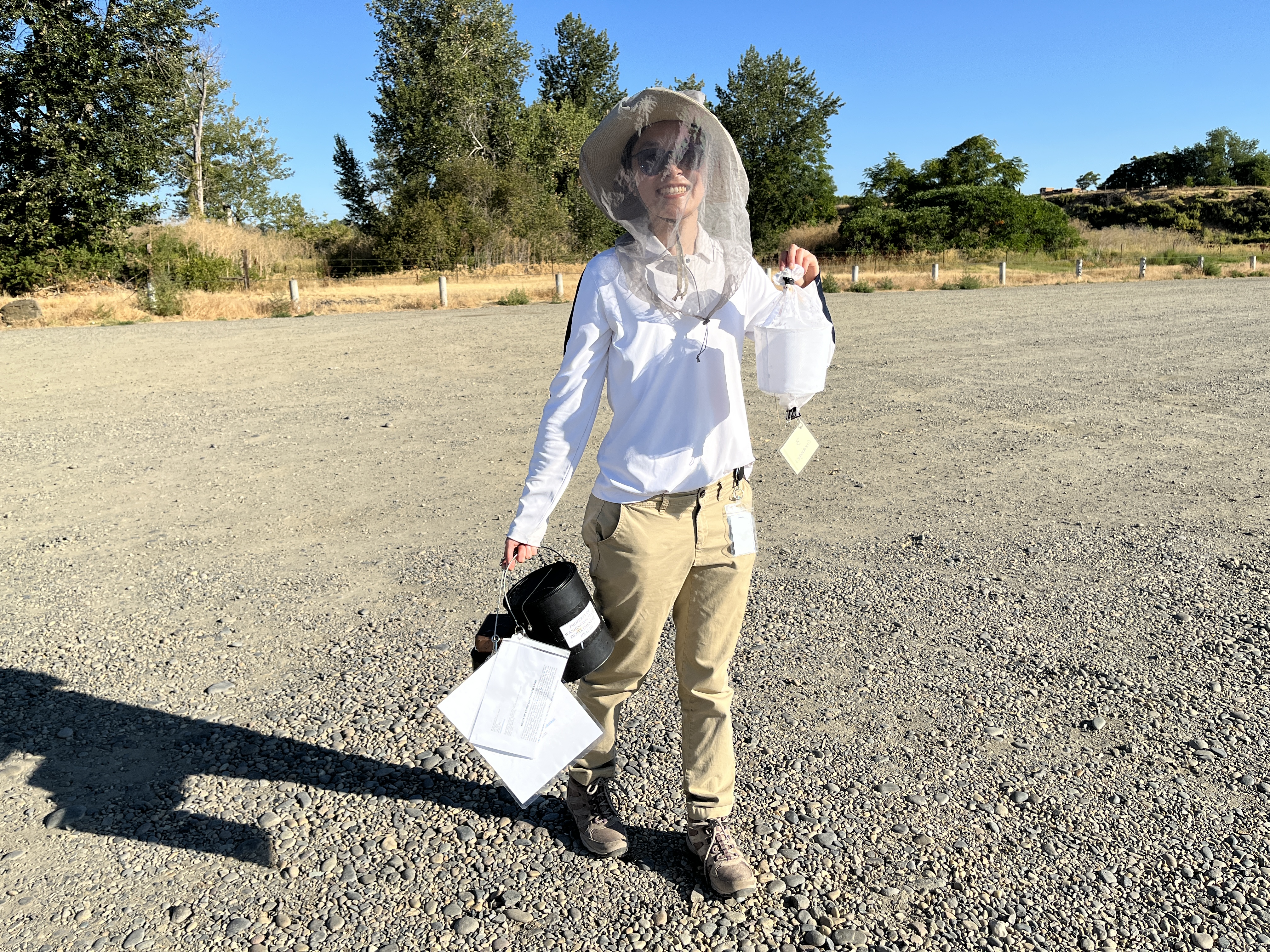 Li, wearing sunglasses, a hat and mosquito net around her face, holds a mosquito trap standing on a gravel surface with trees in the background.