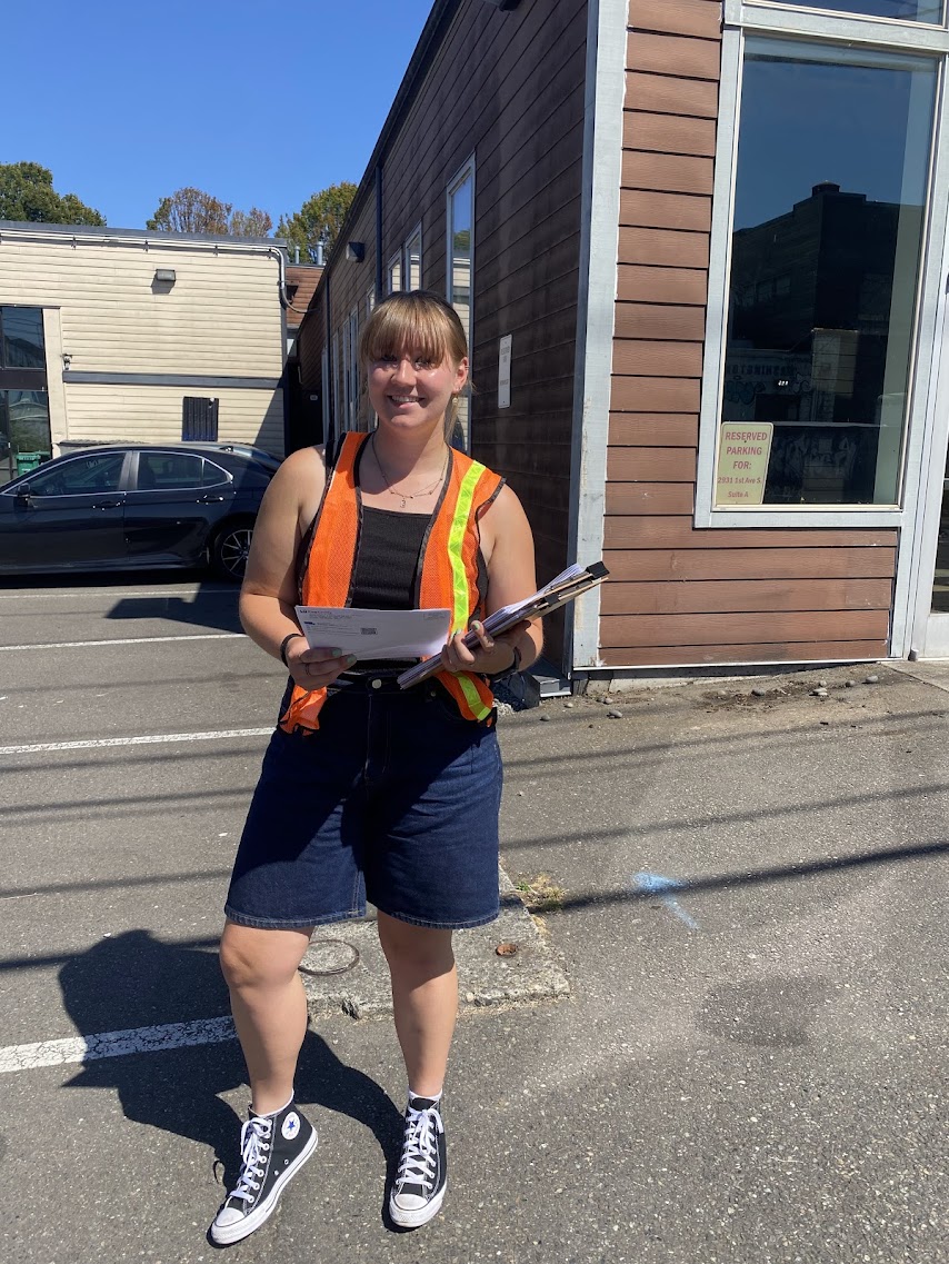 Chamonix Reynolds stands in front of a storefront holding a binder with papers and wearing a safety vest.