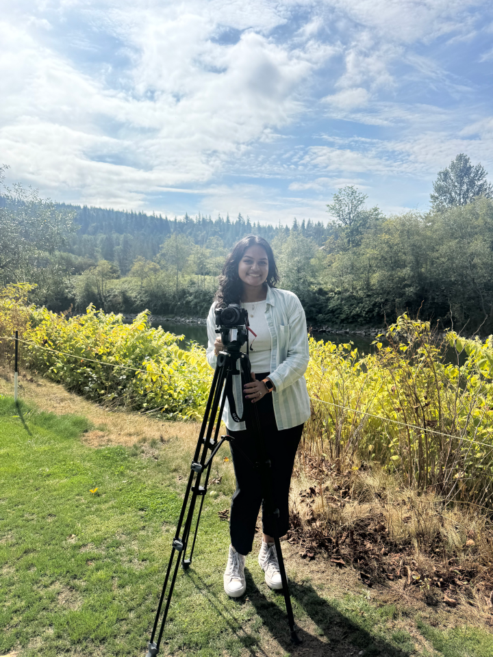 Vijay stands behind a camera on a tripod outside in front of a river and trees.