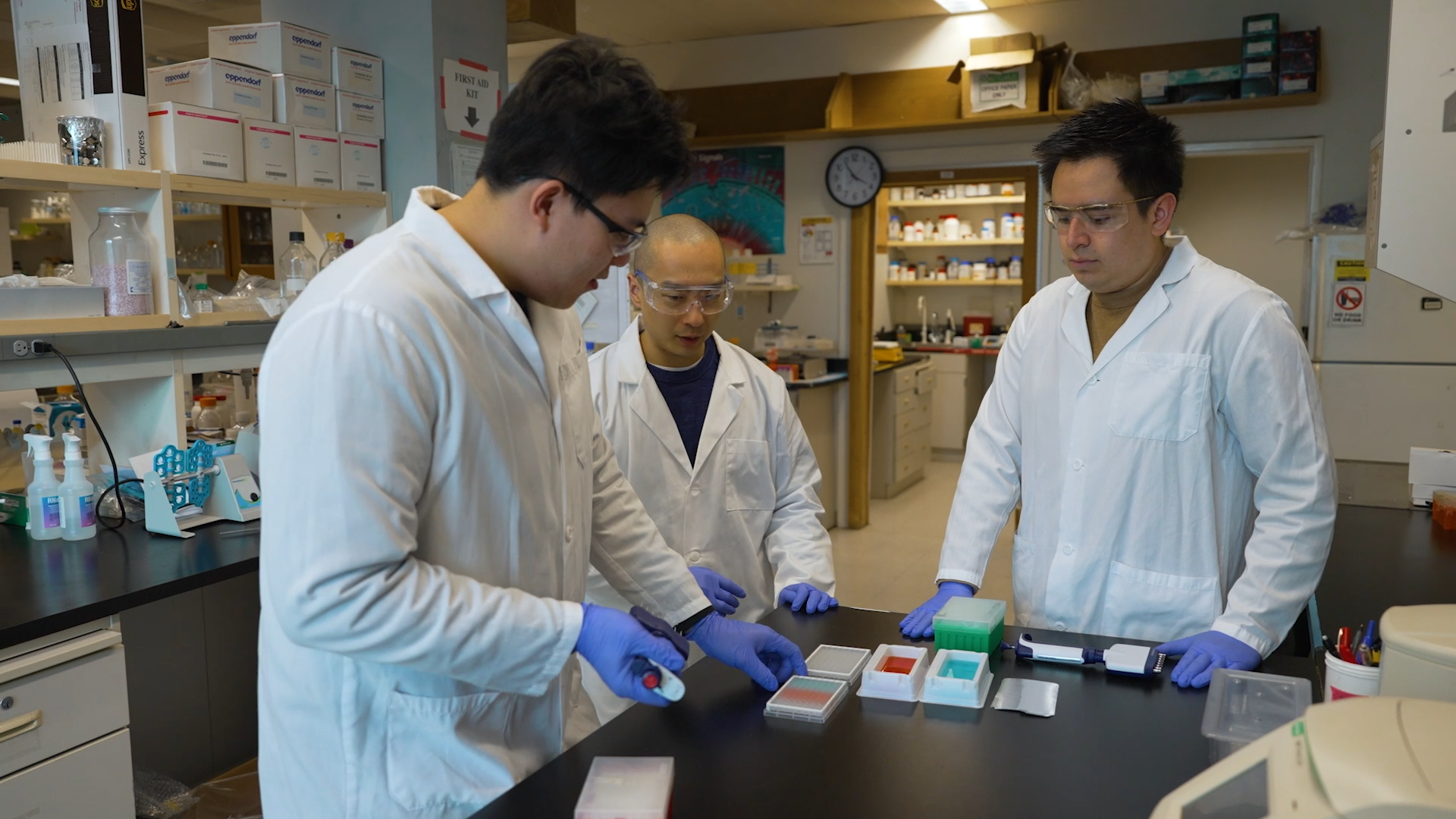 Three men stand around a lab bench looking down at multiwell cell culture plates filled with colorful liquid.