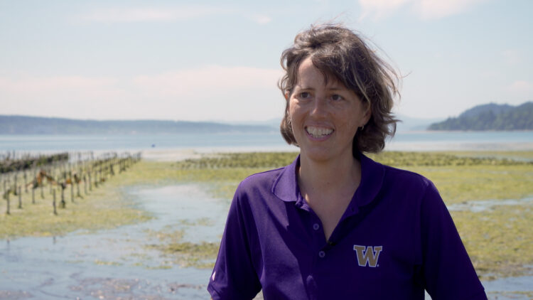 Sarah Collier stands in front of a bay with seaweed floating on the surface and islands in the distance. She is wearing a purple t-shirt with the UW logo on it (a gold W with white outline).