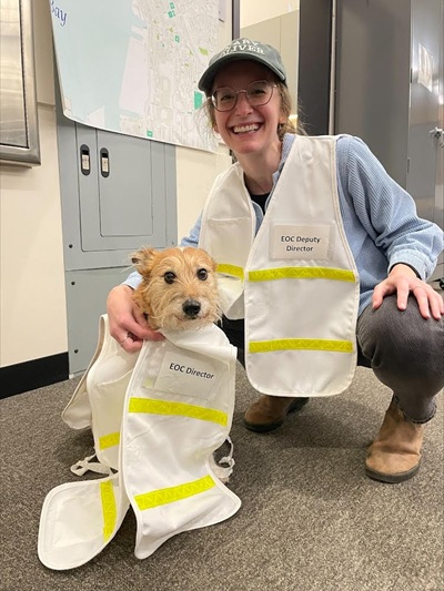 Woman in blue shirt and gray pants wears a baseball cap and white vest with badge saying EOC Deputy Director. She holds a dog who wears a white vest and a badge saying EOC Director.