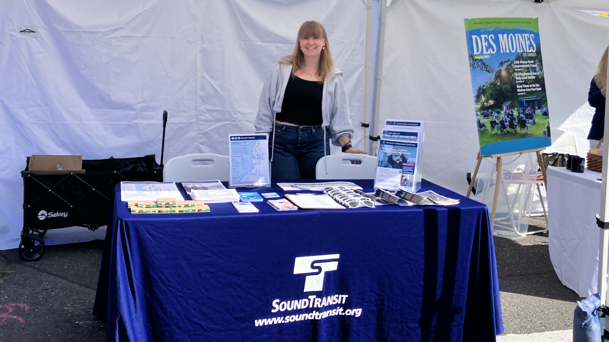 Chamonix Reynolds stands under a canopy, behind a table with a tabletop banner reading "Sound Transit. www.soundtransit.org" On top of the table are informational brochures and promotional swag including sunglasses and notebooks with pens.