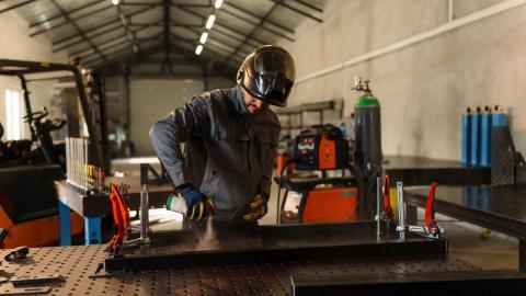 Worker holds spray can over materials on a work bench. They have gloves and a work suit on with a work helmet on their head.