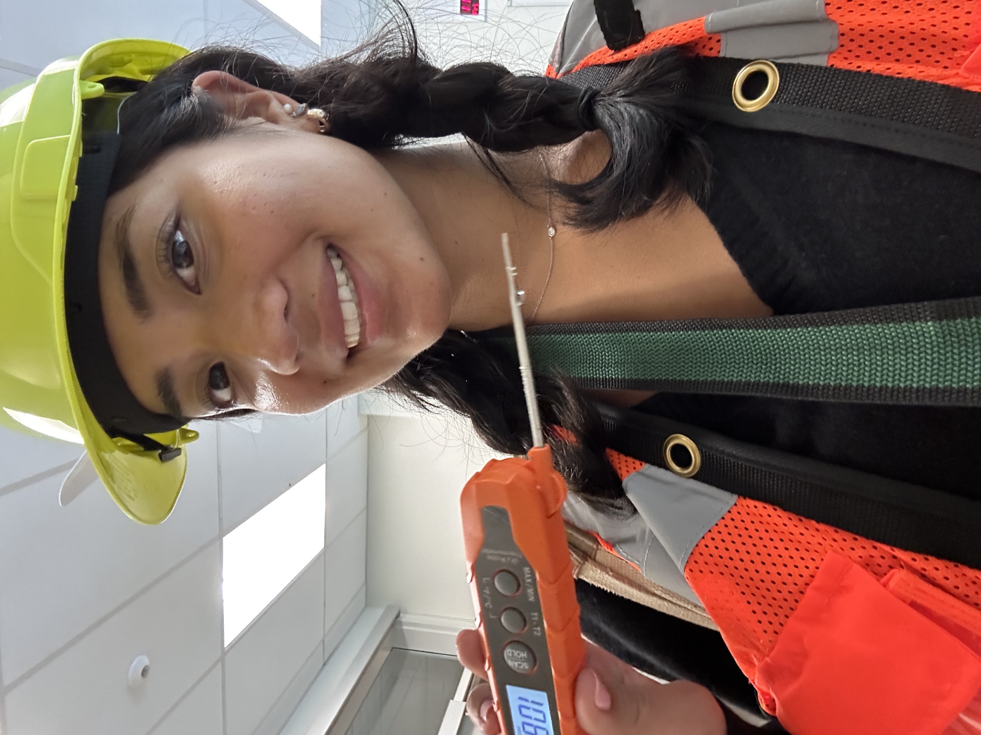 Emily Do smiles wearing a hard hat and safety vest, holding a temperature measuring device.