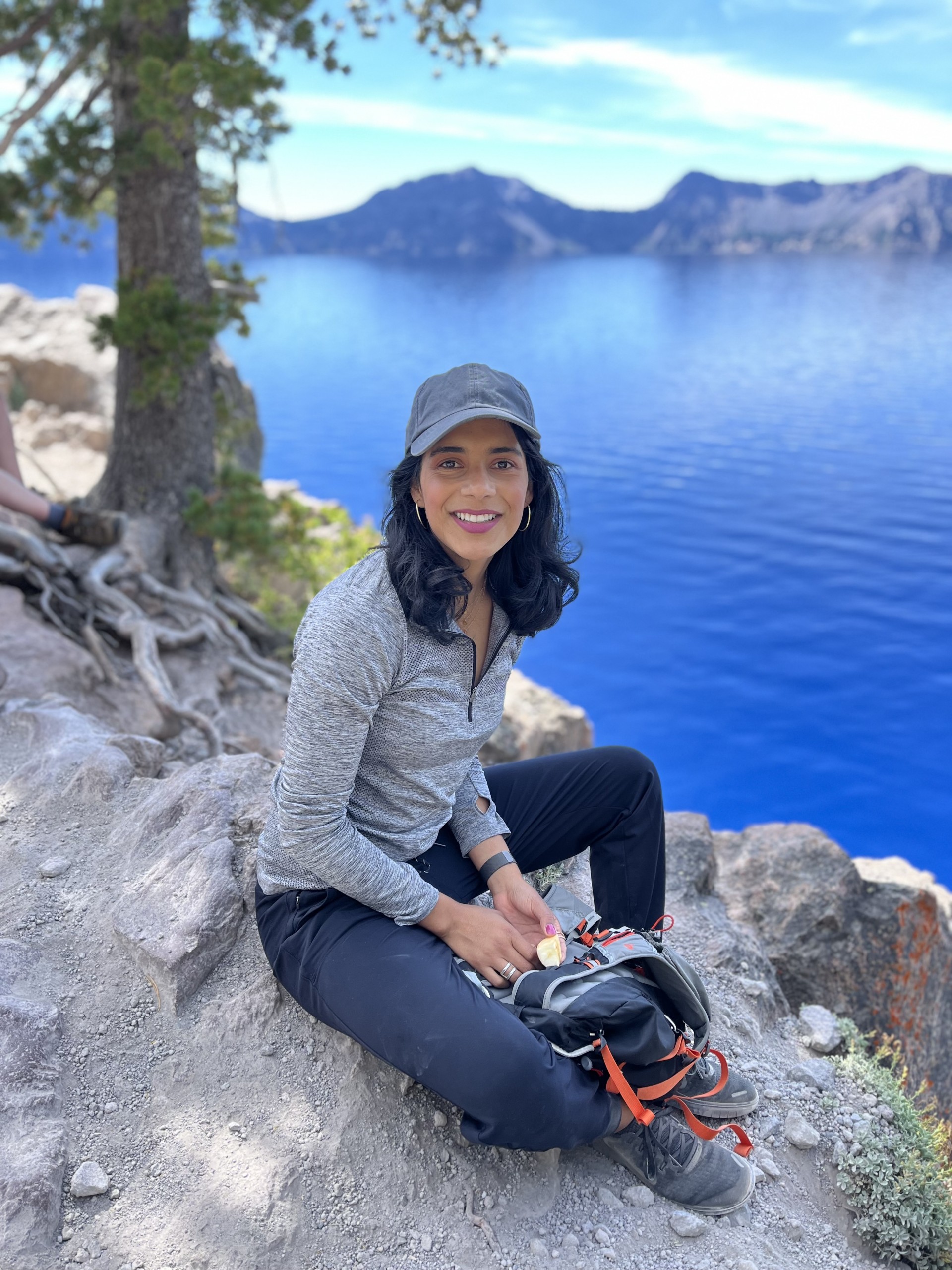 Magali Blanco sits on a cliffside with a tree behind her. A lake and mountains are in the distance behind and below her